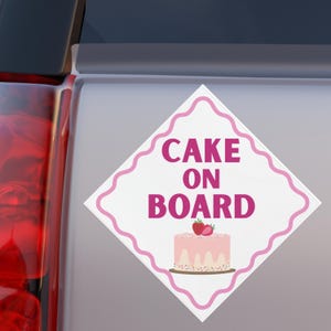 May include: White diamond-shaped sign with pink wavy border and text "CAKE ON BOARD" in pink. Below the text is a pink cake with strawberries on top. The sign is on a light gray surface.