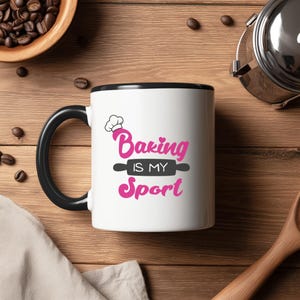 May include: White ceramic mug with a black handle and rim. The mug features the text "Baking IS MY Sport" in pink script and a chef's hat graphic. The mug is on a wooden surface with coffee beans and a wooden spoon.