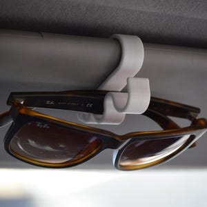 May include: A white plastic sunglasses holder is attached to a car's interior. Brown and tan sunglasses are hanging from the holder. The sunglasses have a rectangular frame and brown lenses. The holder is attached to a grey surface.