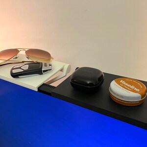 May include: Aviator sunglasses with a gold frame and gradient lenses sit on a white shelf. A black remote and other items are on a black shelf, including a black case and a tin of Vaseline Lip Therapy.