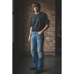 以下が含まれることがあります： 濃いグレーのTシャツと、膝にダメージ加工が施されたライトブルーのジーンズを履いた人。レンガの壁と窓の前に立っています。Tシャツには前面にグラフィックデザインが施されています。