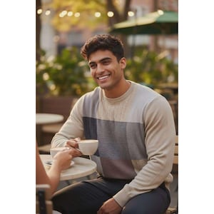 Könnte beinhalten: Ein beigefarbener, grauer und schwarzer Farbblockpullover mit Zopfmuster. Der Pullover wird von einer Person getragen, die an einem Tisch sitzt und eine weiße Kaffeetasse hält. Die Person lächelt, der Hintergrund ist unscharf.