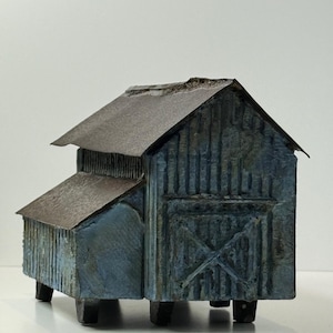 May include: A miniature barn model with a weathered, rustic appearance. The structure features a blue-gray exterior with a brown roof and a small attached shed. The barn is elevated on short, dark legs, giving it a charming, vintage aesthetic.