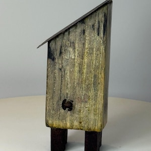 May include: A small, rustic wooden birdhouse with a slanted metal roof. The birdhouse is painted in shades of gray and yellow, with dark brown legs. The design features a small, round opening and a textured surface.