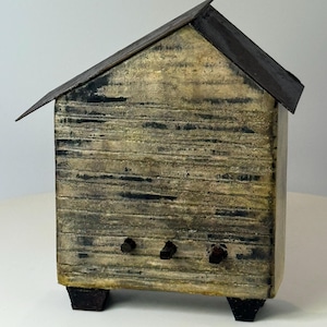 May include: A small, rustic wooden house with a dark metal roof. The house is painted in shades of beige and gray with dark accents. It has three small, dark knobs on the front and sits on four small, dark feet.
