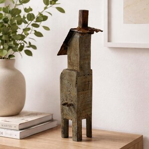 May include: A weathered, abstract sculpture with a rectangular body and a small, square head. The sculpture has a rustic, aged appearance with visible texture and a mix of gray and brown tones. It stands on a wooden surface, suggesting a decorative piece.