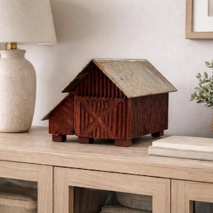 May include: A rustic, wooden barn model with a weathered metal roof sits on a light-colored cabinet. The miniature barn is painted a deep reddish-brown and features a cross-shaped design on the door. A lamp and a plant in a woven basket are also visible.
