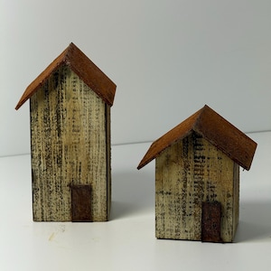 May include: Two rustic wooden miniature houses with brown rusted metal roofs and small doors. The houses have a weathered, distressed appearance with vertical wood grain and a light, neutral color palette.
