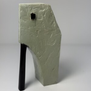 May include: A unique, abstract sculpture in a muted green hue. The textured surface contrasts with a sleek, black, curved support. A small, square, black detail is embedded in the top. The sculpture's design is modern and artistic.