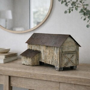 May include: A rustic, weathered barn model with a metal roof and a small attached shed. The barn is painted in shades of white and gray, with visible wood grain. It sits on a wooden table, next to a white vase and a stack of books.