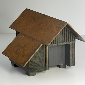 May include: A rustic, miniature wooden barn sculpture with a brown, angled roof and dark gray walls. The barn has a small opening and sits on four rectangular feet. The wood has a textured, weathered appearance.