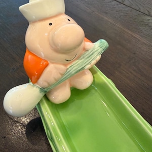 May include: A whimsical ceramic serving dish featuring a sailor figure with a white hat and orange vest. The figure holds a green celery stalk, resting on a bright green rectangular dish. The dish is ideal for serving snacks or appetizers.