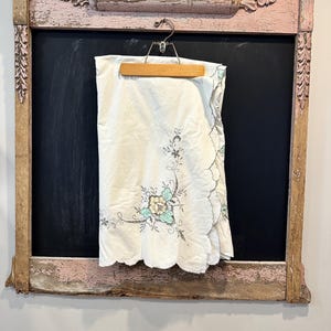 May include: A white, scalloped-edge tablecloth with floral embroidery in shades of blue, yellow, and gray, hanging on a wooden hanger. The tablecloth is displayed against a black background within a distressed, pink-trimmed frame.