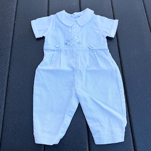 May include: Light blue and white striped baby romper with a white collar and short sleeves. The romper features a small embroidered design on the chest and button closures. The fabric has a subtle vertical stripe pattern.
