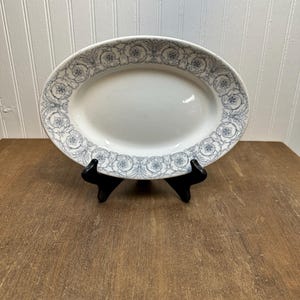 May include: Oval white serving platter with a blue floral design around the rim. The platter is displayed on a black stand against a wooden surface and a white paneled wall.