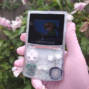 Puede incluir: Consola portátil Game Boy Color transparente que muestra una escena de videojuego. La carcasa transparente revela los componentes internos. La pantalla muestra un personaje con ropa verde. El texto "GAME BOY COLOR" es visible.