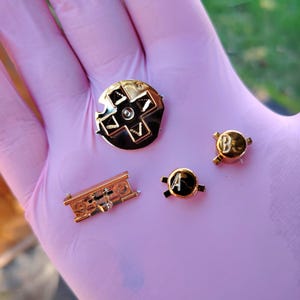 May include: A selection of gold-coloured gaming controller components, including a directional pad, a rectangular piece, and two buttons marked "A" and "B". The parts are presented on a pink surface.