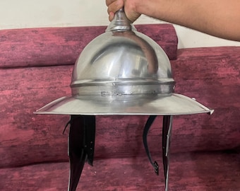 May include: A silver-colored metal helmet with a wide brim and black cheek guards. The helmet is held by a hand, showcasing its polished surface and historical design. The helmet's design suggests a Roman or medieval influence.