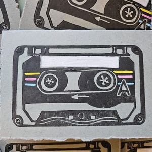 May include: A gray cassette tape print with black details and colorful stripes. The cassette tape design includes the letter "A" and star-like symbols. The image is a print on a textured, light gray paper.