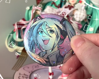 May include: A round, holographic button featuring a smiling anime character with light blue hair and a black headset. The button is held in front of a figurine with long, light green hair and a white dress. The figurine is on a pink base.