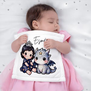 Peut inclure: Une couverture blanche pour bébé avec une princesse et un dragon de dessin animé. La princesse porte une couronne et une robe bleue à étoiles. Le dragon est gris et bleu. La couverture porte le nom "Esmi" en écriture noire.