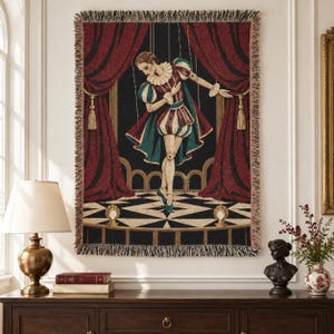 May include: A woven tapestry throw blanket featuring a marionette puppet dressed in a striped doublet and breeches, performing on a stage with dark red curtains, a black and white checkered floor, and visible strings against a dark background.