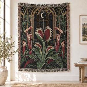 May include: A woven throw blanket featuring a dark botanical scene with a central Venus flytrap and pitcher plants. The background displays a gothic arched window under a crescent moon and stars. The blanket has a fringed edge on all sides.