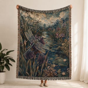 May include: A woven tapestry throw blanket featuring a detailed nature scene with a large dragonfly in the foreground, purple iris flowers, lily pads on a pond, and a forest landscape in the background, finished with a decorative fringe border.