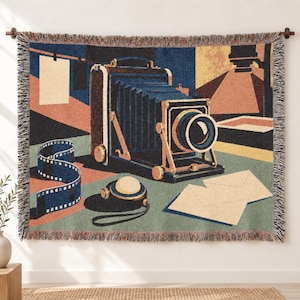 May include: A woven wall tapestry featuring a vintage bellows camera, a strip of 35mm film, a round light meter, and two envelopes. The scene is rendered in a palette of navy blue, muted orange, cream, and sage green with a fringed edge finish.