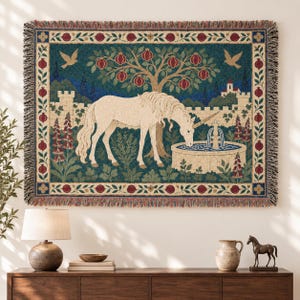 May include: A rectangular woven tapestry featuring a cream-colored unicorn drinking from a stone fountain. The scene includes a pomegranate tree, birds, and a castle wall, all framed by a decorative border of pomegranates and leaves with fringed edges.