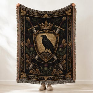 May include: A woven throw blanket featuring a black background with a central gold shield displaying a black raven. The design includes a gold crown above the shield, two crossed swords behind it, and decorative borders with oak leaves and thistles.