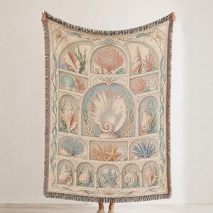 May include: A woven throw blanket featuring a detailed tapestry design of various seashells, coral, and seaweed arranged within arched architectural compartments. The blanket has a fringed edge and a color palette of soft beige, blue, and pink.