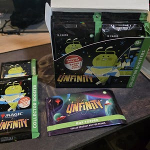 May include: A Magic: The Gathering Unfinity Collector Booster display box containing multiple sealed card packs, with two additional individual booster packs and one Unfinity Box Topper pack featuring space-themed graphics and green accents.