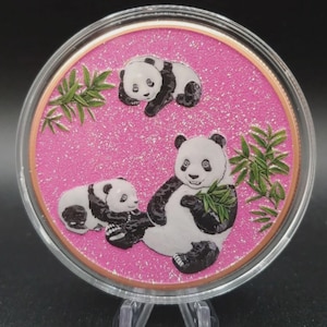 May include: A circular commemorative coin featuring a vibrant pink glitter background. The design depicts three black and white pandas in various poses, with one adult panda holding green bamboo leaves and two smaller pandas nearby.