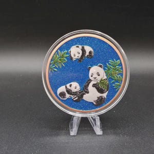 May include: A circular decorative piece featuring a glittery blue background with three black and white panda bears and green bamboo leaves. The design is encased in a clear, round frame with a copper-colored border, supported by a clear stand.