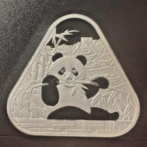 May include: A triangular silver-toned coin or medallion featuring a relief design of a panda sitting and eating a bamboo stalk. The background depicts a bamboo forest and a section of the Great Wall of China, all enclosed within a raised border.