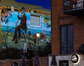 Puede incluir: Una vista exterior de un edificio al anochecer. Un mural representa a un hombre en una bicicleta antigua. El edificio es de ladrillo con un balcón y un letrero que dice "Cellar". Hay sombrillas cerradas y una valla de metal negro.