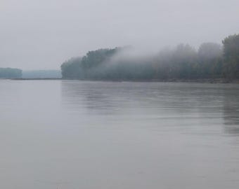 Può includere: Un paesaggio sereno con un ampio fiume calmo sotto un cielo nuvoloso e coperto. La riva lontana è fiancheggiata da una fitta foresta, parzialmente oscurata da uno strato di nebbia o foschia, creando una tavolozza di colori tenui e smorzati.