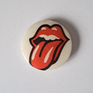 Peut inclure: Un bouton rond blanc cassé avec le célèbre logo des lèvres et de la langue rouges, bordé de noir. Le design est centré sur le bouton, qui présente une légère patine.