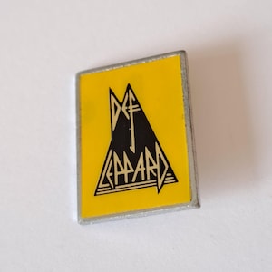 Könnte beinhalten: Eine rechteckige Anstecknadel mit gelbem Hintergrund und einem schwarzen Dreieck-Design. Die Anstecknadel zeigt den Bandnamen "Def Leppard" in einer stilisierten Schriftart. Die Anstecknadel hat einen silberfarbenen Rand.