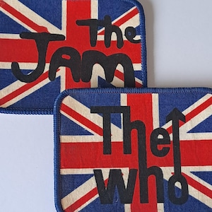 May include: Two rectangular patches featuring the Union Jack flag design. One patch displays the text "The Jam" in black, while the other reads "The Who" with an upward arrow. The patches have a blue background with red and white stripes.