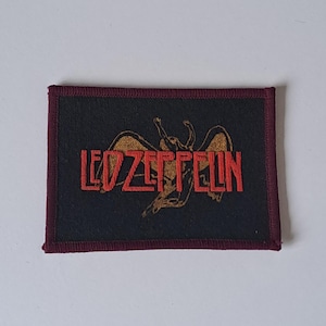 May include: A rectangular patch featuring the Led Zeppelin logo. The patch has a black background with the band's name in red block letters and a gold illustration of a winged figure. The border is a dark burgundy color.