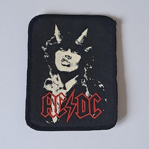 Könnte beinhalten: Schwarzer Aufnäher mit einem stilisierten Bild einer Person mit Hörnern und dem Bandnamen "AC/DC" in Rot. Der Aufnäher hat eine rechteckige Form mit abgerundeten Ecken und einem genähten Rand.