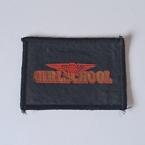 Puede incluir: Un parche rectangular negro con la inscripción dorada "GIRLSCHOOL". Encima del texto hay un emblema rojo con un diseño alado. El parche tiene un borde negro. Probablemente un emblema escolar.