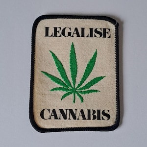 Puede incluir: Un parche rectangular con fondo beige y borde negro. El parche presenta las palabras "LEGALISE CANNABIS" en letras negras, con una hoja de cannabis verde en el centro.