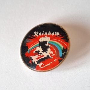 Può includere: Spilla ovale con sfondo nero e la scritta "Rainbow" in corsivo bianco. La spilla presenta un pugno, un arcobaleno e accenti rossi e arancioni. La spilla ha un bordo in metallo color argento.