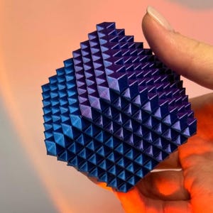 Könnte beinhalten: Eine geometrische, dreidimensionale Skulptur mit strukturierter Oberfläche. Das Objekt hat eine würfelartige Form, die aus zahlreichen kleinen, dreieckigen Pyramiden besteht. Die Farbe wechselt von Blau zu Lila und erzeugt einen Farbverlauf. Die Skulptur wird in einer Hand gehalten.