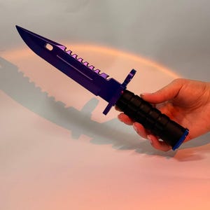 Puede incluir: Un cuchillo morado y negro con borde dentado y mango negro. El cuchillo se sostiene en una mano sobre un fondo blanco. La hoja tiene punta y un agujero cerca del mango.