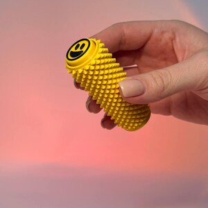 Könnte beinhalten: Ein leuchtend gelbes, zylindrisches sensorisches Werkzeug mit einer texturierten, stacheligen Oberfläche. Das obere Ende verfügt über eine flache, kreisförmige gelbe Kappe mit einem schwarzen Smiley-Symbol. Das Objekt hat ein gleichmäßiges Muster.