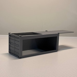 Könnte beinhalten: Ein grauer Miniatur-Seecontainer mit offenem Deckel. Der Container hat eine rechteckige Form mit geriffelten Seiten und einem Klappdeckel. Der Innenraum ist hohl und steht auf einer strukturierten Oberfläche.